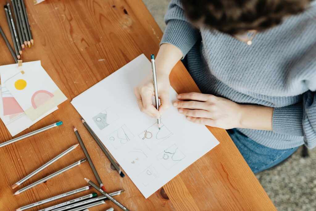 Artist sketching designs on paper with pencils on a wooden desk from above.